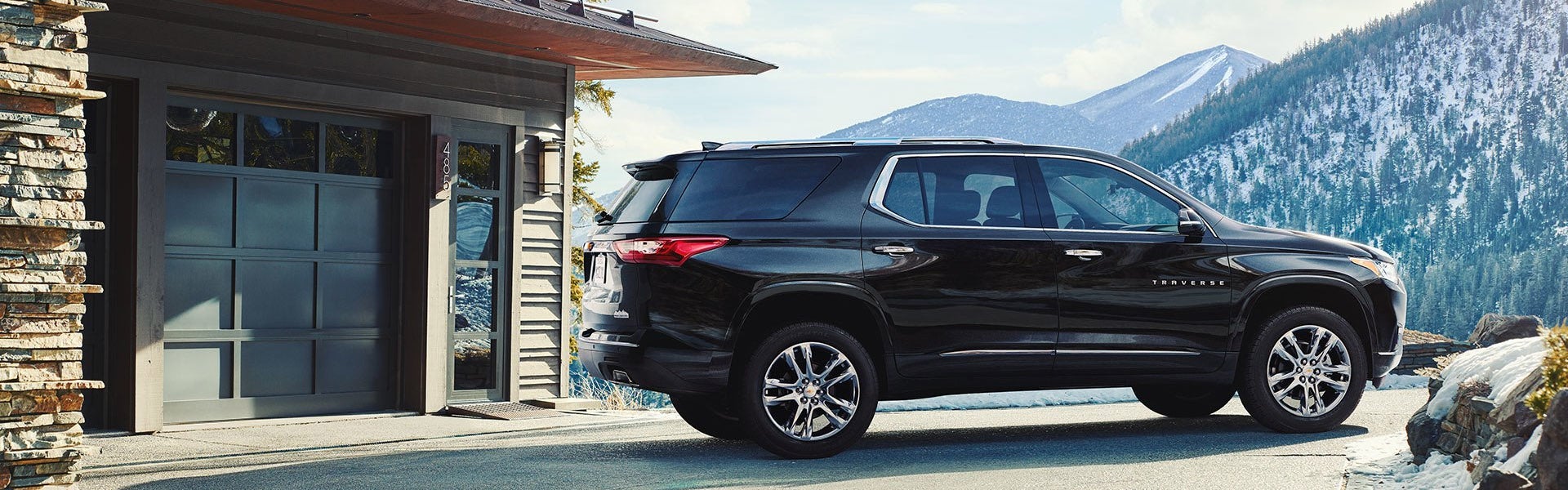 Pre-owned black Chevrolet Traverse SUV parked in front of a home garage with snowy mountains behind it.