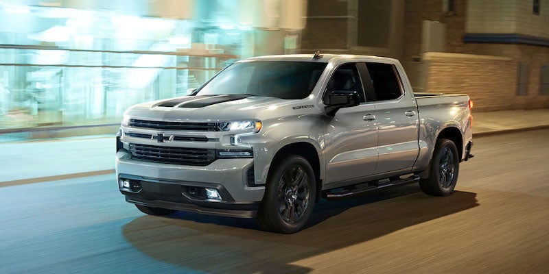 A silver Chevrolet Silverado pickup truck driving through a city street at night.